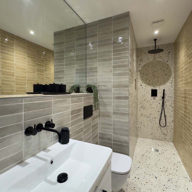 Wet room-style shower room with grey and peach tiles, wall-mounted basin and toilet, black taps and shower head, and a wall-to-wall mirror in a contemporary Sneyd Park home by Ivywell Interiors