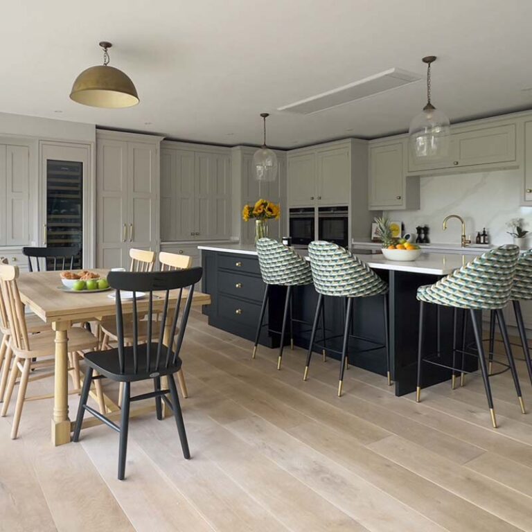 Open-plan bespoke kitchen with shaker-style panels, large dark blue island, colourful upholstered stools, and a light oak dining table for six in a contemporary Sneyd Park home by Ivywell Interiors